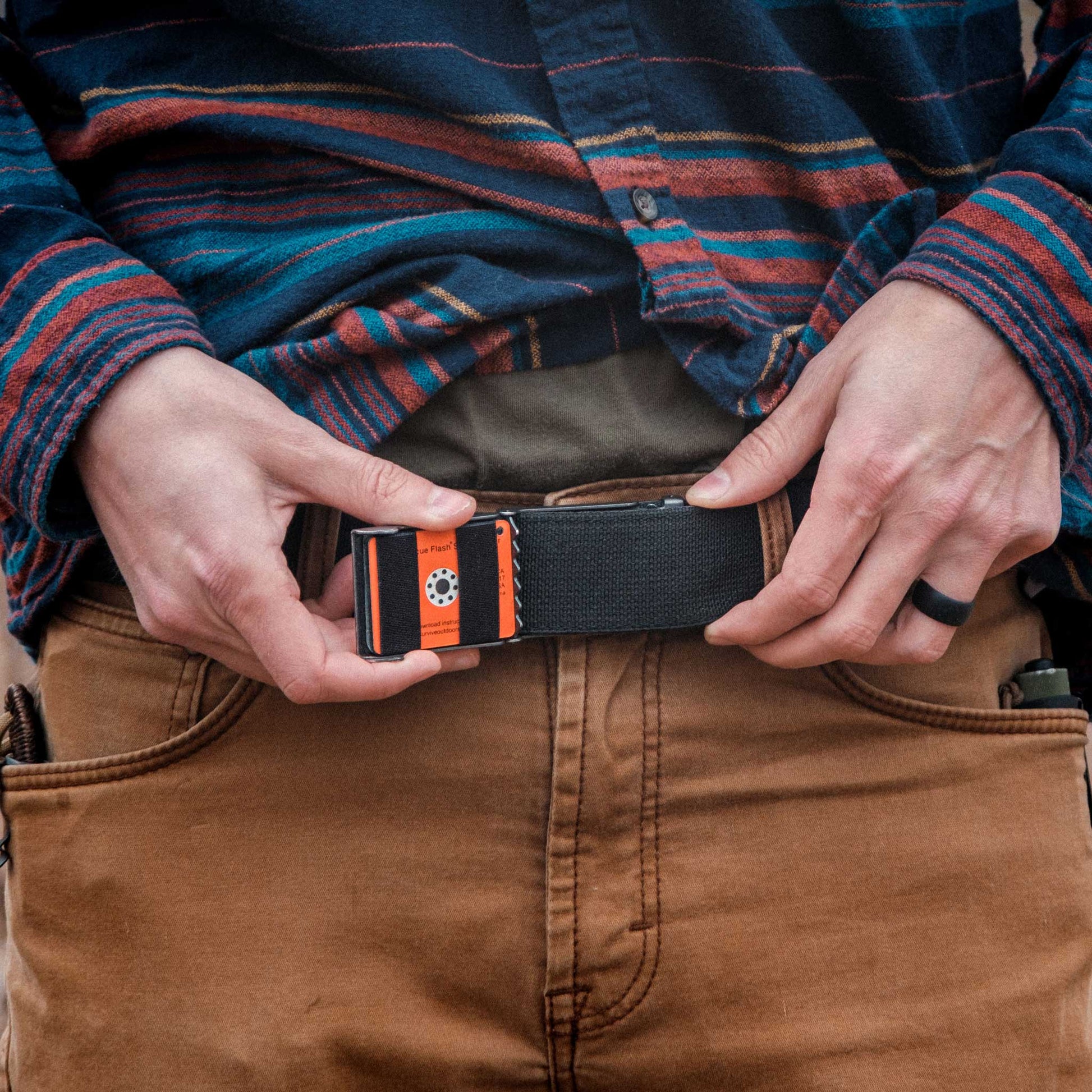 Person holding a black belt with orange accents, wearing a plaid shirt and brown pants.