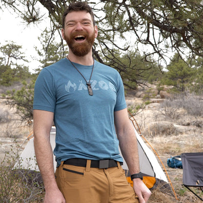 Man in a blue t-shirt and brown pants standing outdoors with camping gear in the background